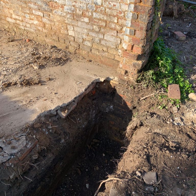 Excavated trench next to a brick wall, revealing soil and debris.