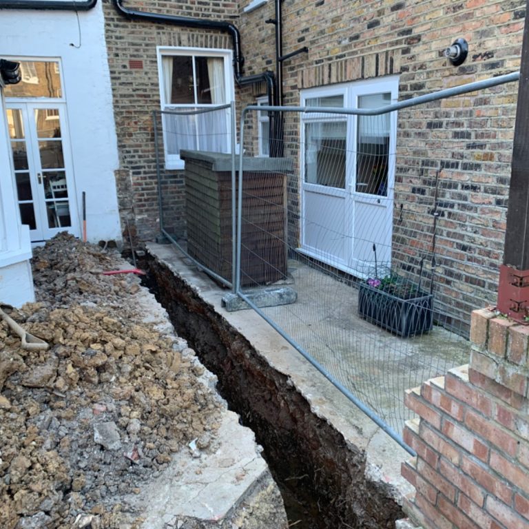 Excavated area near brick wall, showing a deep trench beside a house foundation.