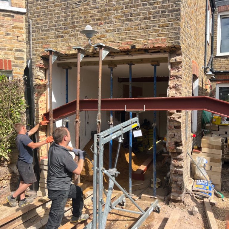 Men supporting a steel beam during home renovation, with construction materials around.