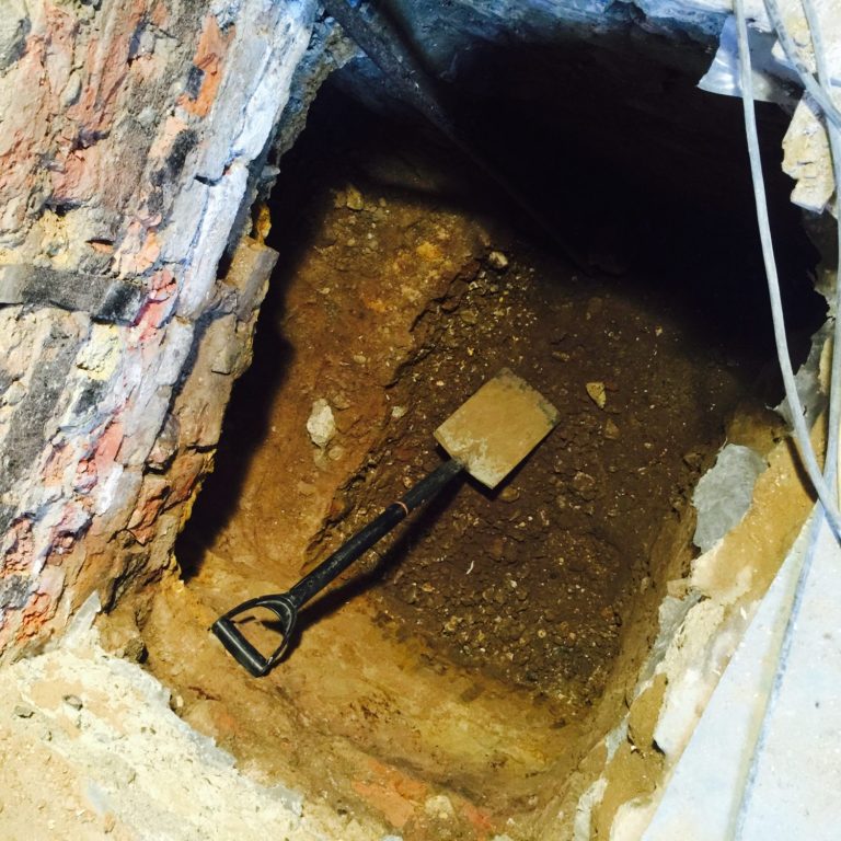 An excavation hole for underpinning a party wall with a shovel resting in the dirt.